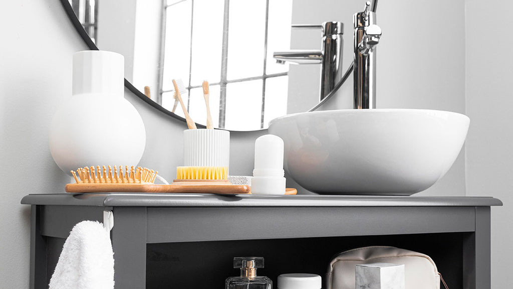 A modern bathroom with grey tiles, a sleek white sink, mirror, and bathroom shelf with neatly folded towels, toothbrush, soap, and a white backpack on the ground.