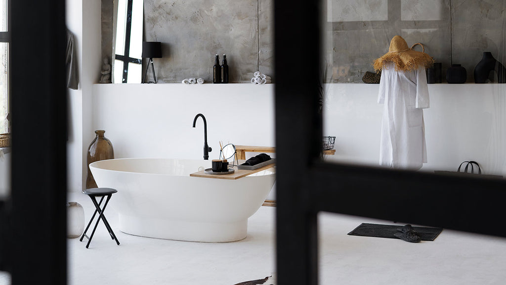 A spacious black and white bathroom with a white bathtub, black folding stool, white mannequin with a straw hat, black slippers, white bowl, and a mirror with a cross in the middle.