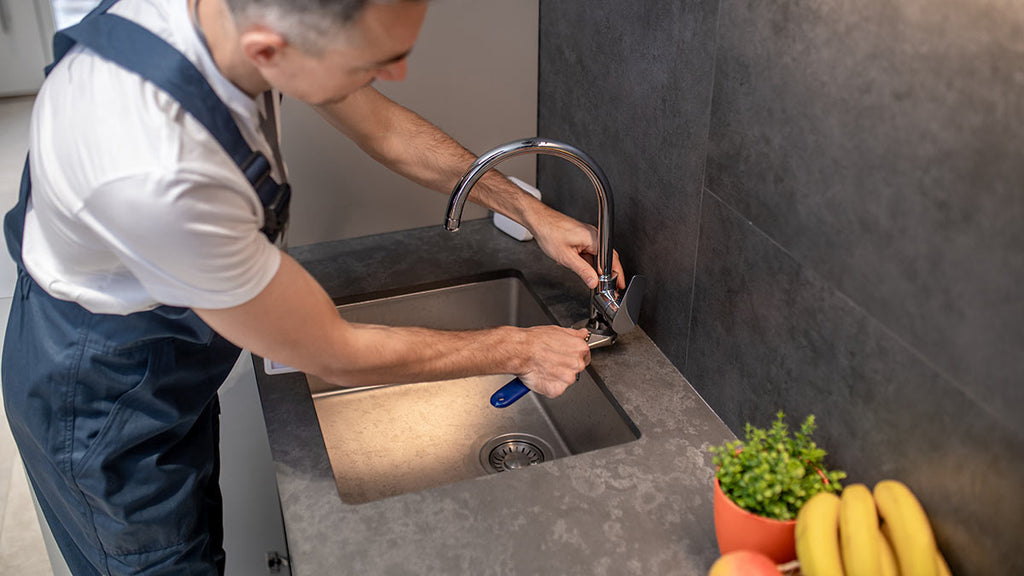 Professional plumber repairing sink and faucet in kitchen, woman preparing food, basket with ripe bananas.