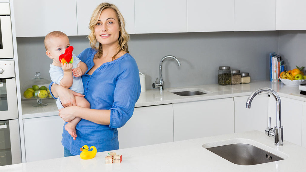 Woman in blue apron holding baby and cleaning kitchen sink, stainless steel faucet, yellow rubber duck.