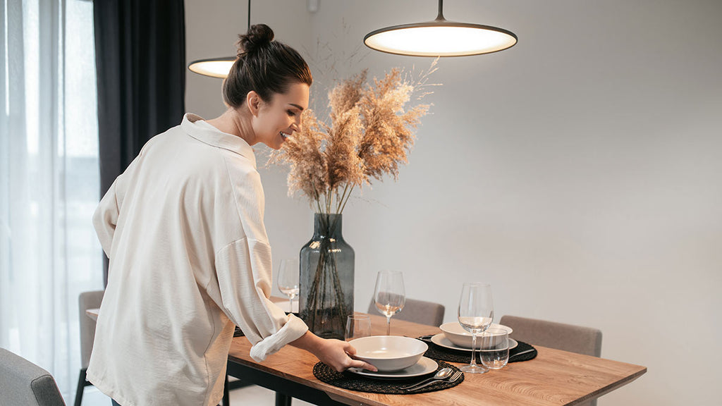 woman in a white shirt sets the table in a cosy dining room, illuminated by a pendant light. A stove and a wine glass add to the room's warm ambiance.