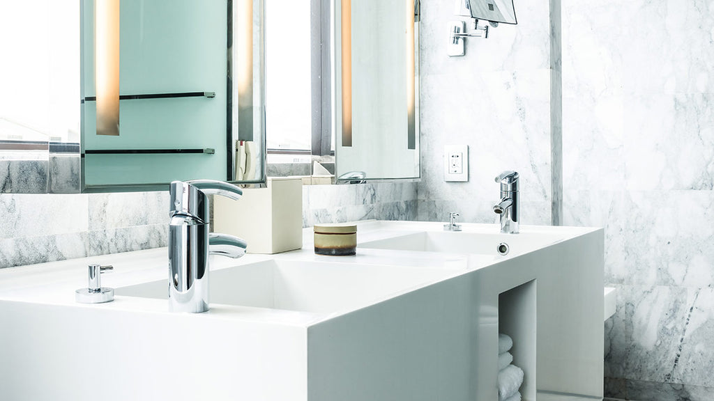 Well-lit bathroom with white tiles, large rectangular mirror, chrome faucets, marble counter, white basin, soap dispenser, small jar, and secondary mirror.