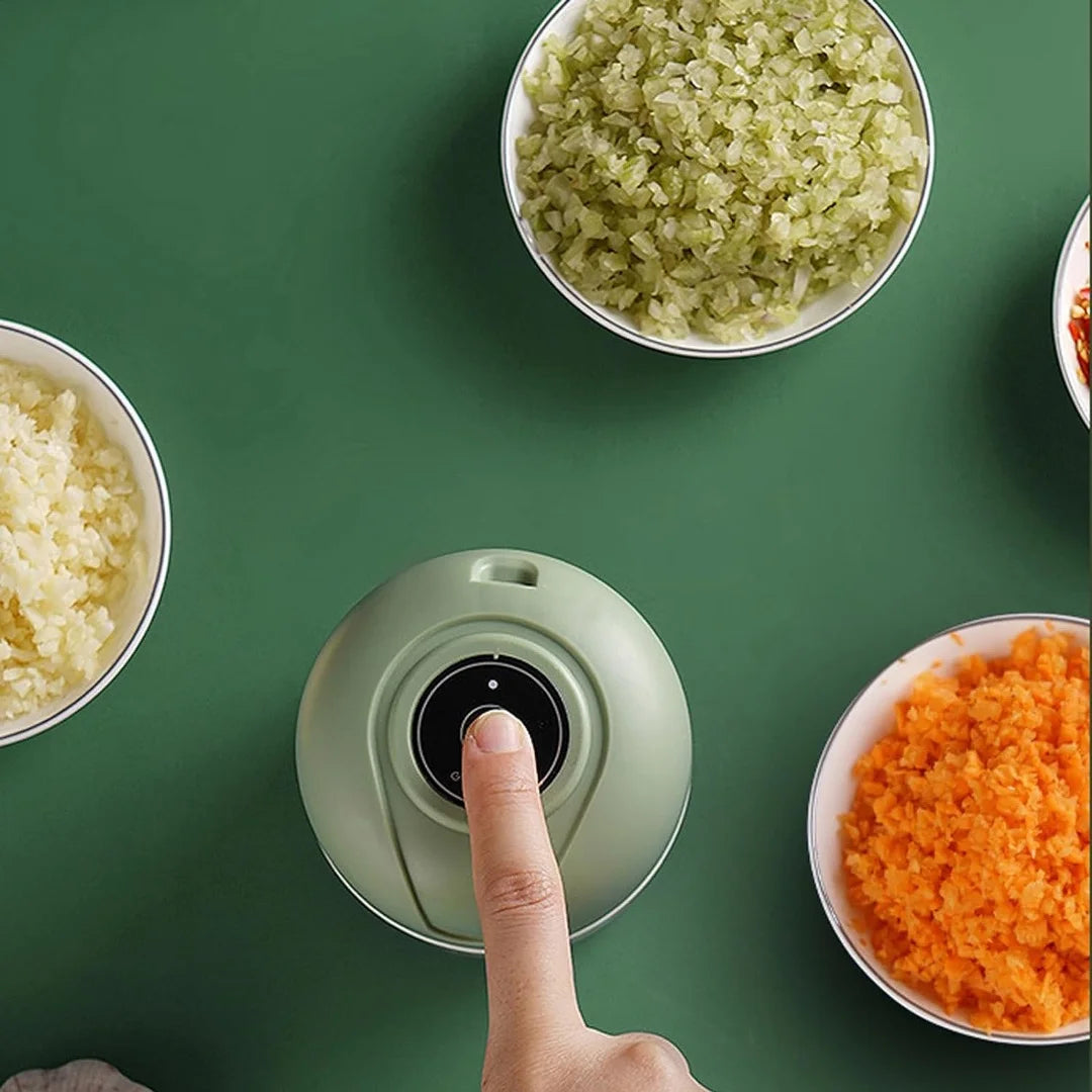 A finger pressing a button on a green kitchen gadget surrounded by bowls of chopped vegetables on a green surface, showcasing the Electric Garlic Chopper - Portable USB Food Processor for Kitchen Gadget by The Stuff Box, made from durable materials.