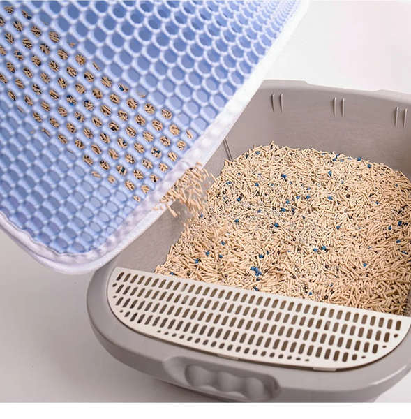 A blue, honeycomb-designed *Waterproof Cat Litter Mat* by *The Stuff Box*, featuring a double layer design and made of cat-friendly material, is being lifted to pour light brown, granulated material into a gray litter box with a grate.