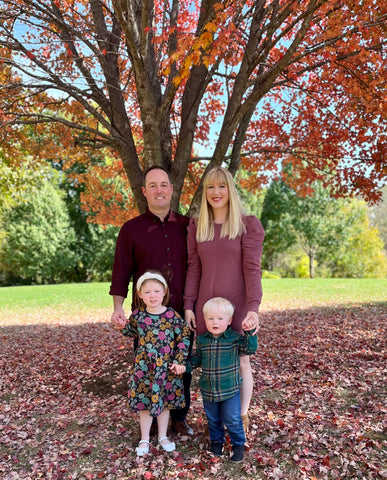 Husband, Wife and two small children standing in front on a tress with leaves on the ground. 