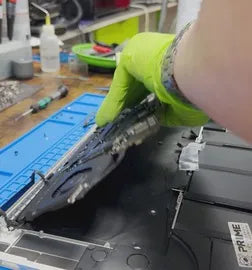 Un técnico de Prime Tech Support en Miami, Florida,<!--nl--> con guantes verdes, trabaja en el reensamblaje de la placa lógica y los ventiladores de<!--nl--> MacBook Pro sobre una alfombra de trabajo azul con herramientas y suministros alrededor