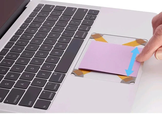 Close-up view of a laptop keyboard with a pink note being inserted onto the touchpad area, demonstrating how to level the Trackpad