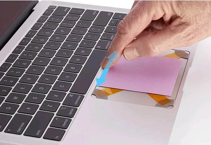 Close-up view of a laptop keyboard with a pink note being inserted onto the touchpad area, demonstrating how to level the Trackpad