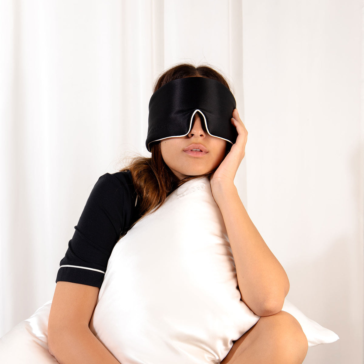 Model with black Drowsy sleep mask covering eyes hugging a white silk pillow with a white silk backdrop