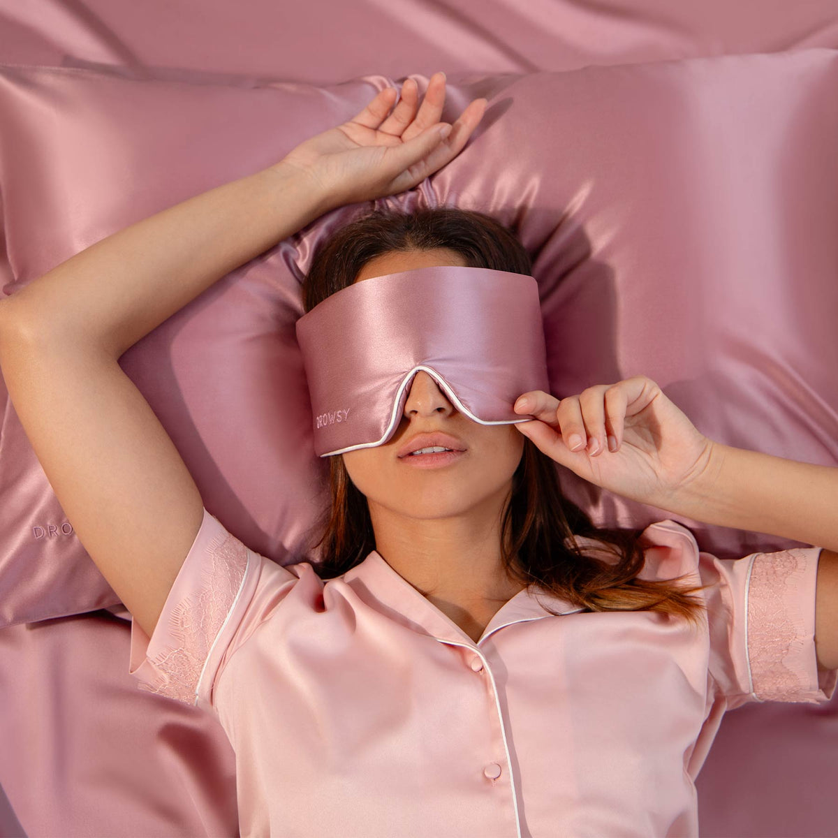 Model with pink rose coloured Drowsy sleep mask covering eyes on pink silk sheets