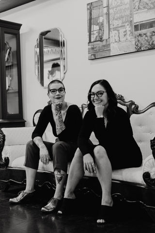 Black and white photo of co owners Nikki and Nicole sitting on a couch in their shop smiling