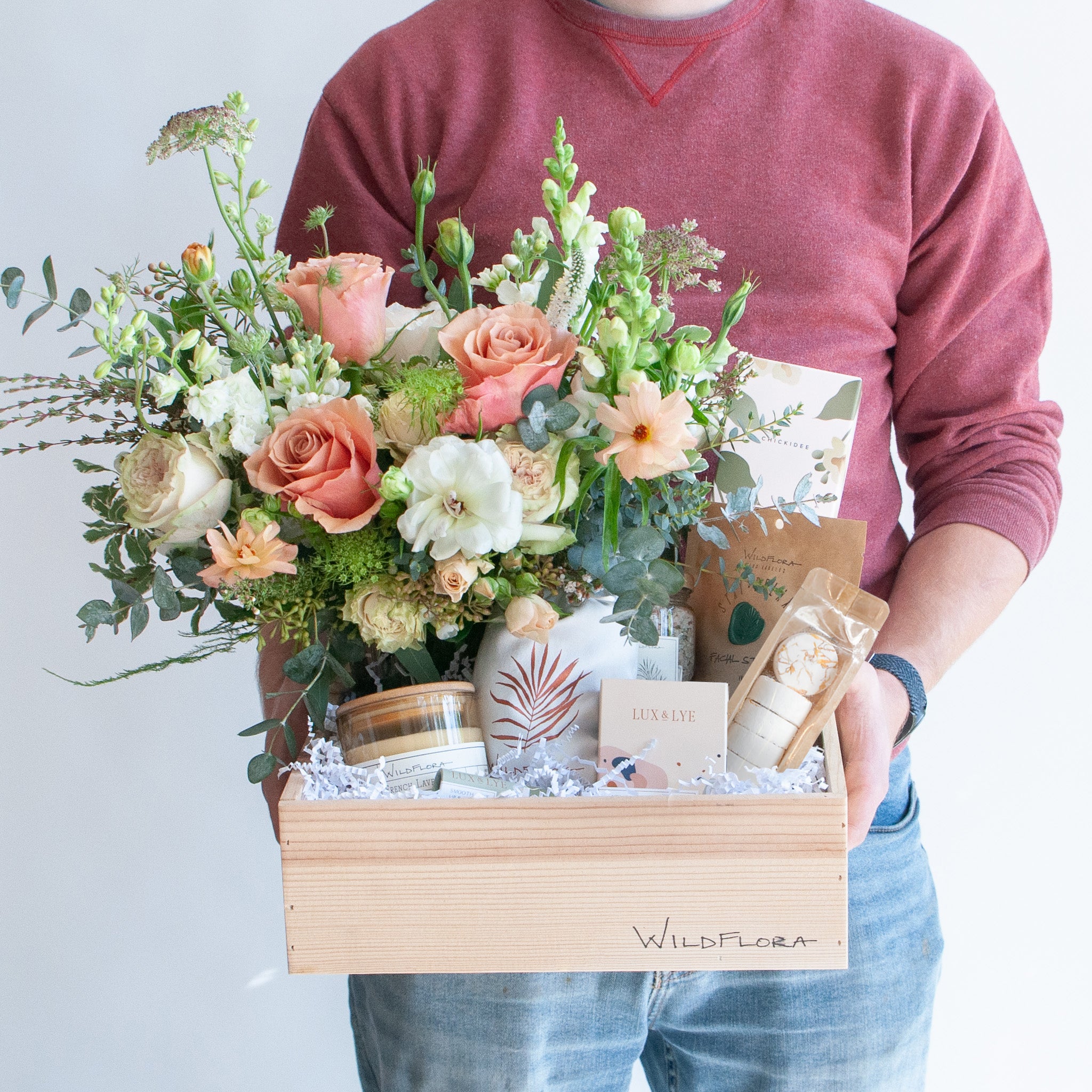 A man holding a wood gift box. In it is a white, chartreuse, peach, and salmon-colored flower arrangement, including rose, veronica, anemone, eucalyptus, queen anne's lace, and snapdragon. It also includes a candle, shower steamers, a lavender sachet, chapstick, bath salts, a facial steam, a reed diffuser and soap.