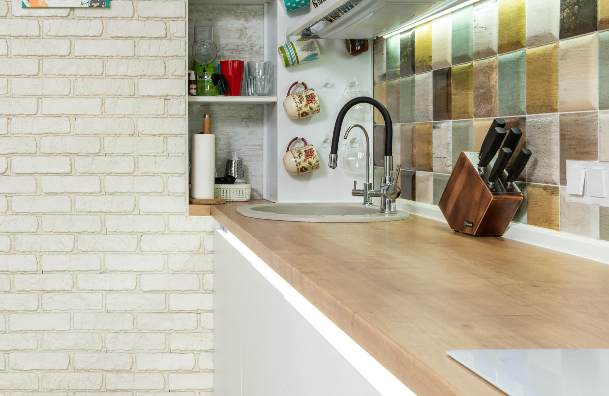 Cozy kitchen corner with a wooden countertop, brick wall, and a colorful, rustic tile backsplash, featuring a modern sink and knife block.