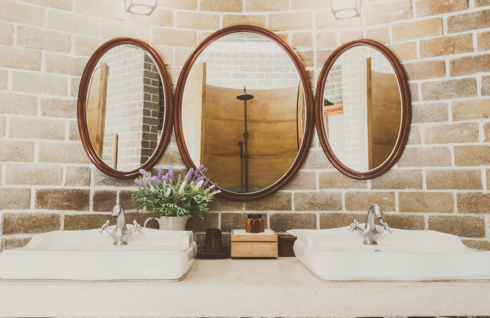 Rustic bathroom with dual white basins, vintage faucets, and three oval mirrors set against a warm brick tile backdrop.