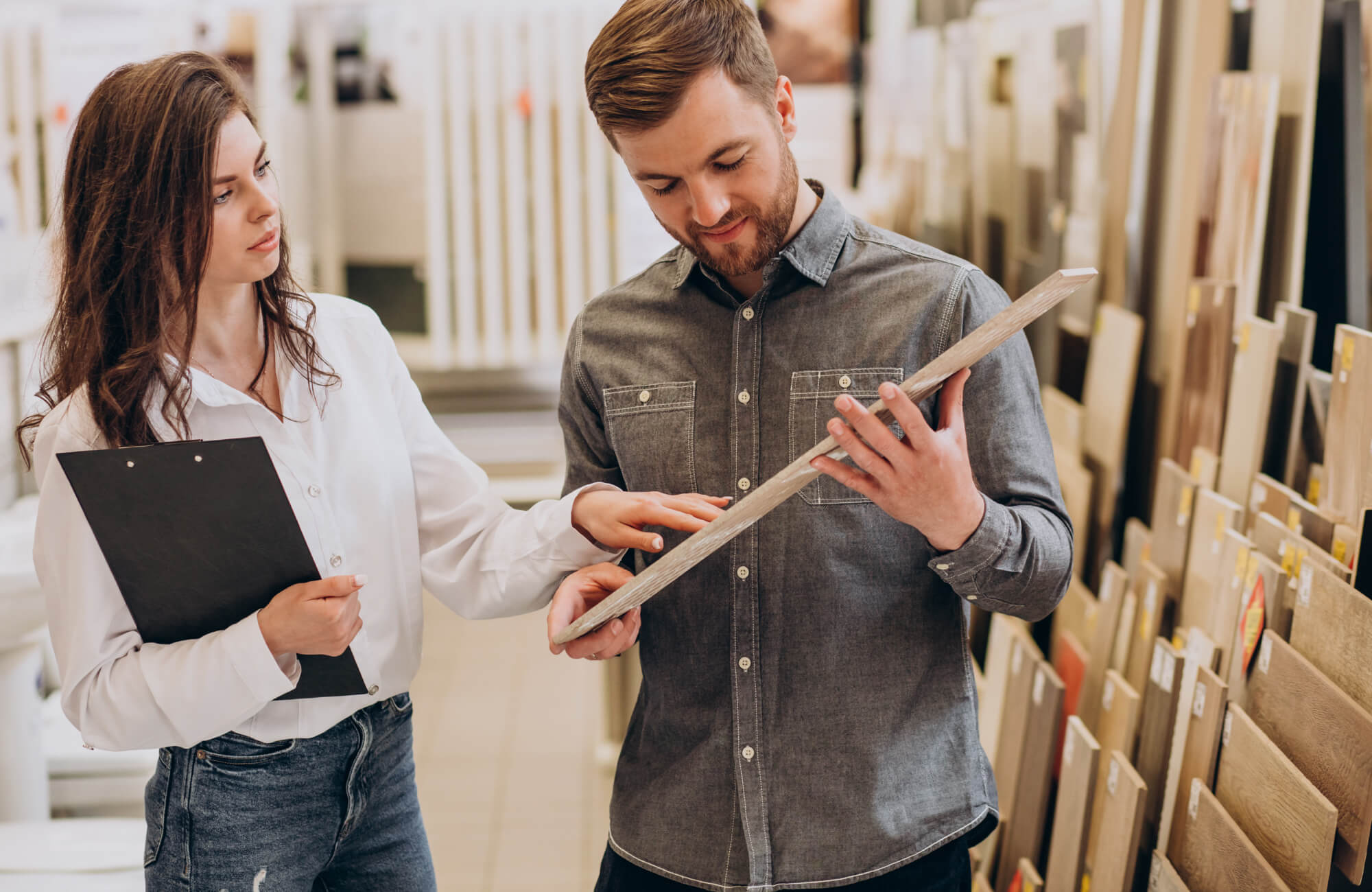 A man examines a wood-look tile sample at a flooring showroom, while a sales lady discusses texture and design options to find the perfect fit for their home renovation.