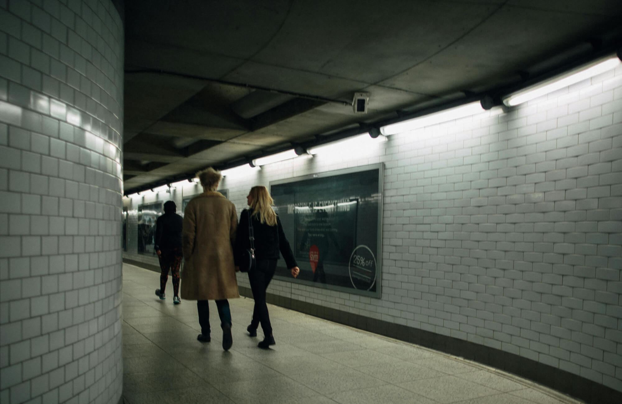 Dimly lit subway tunnel with white subway tiles along the curved walls, as three people walk toward the exit under fluorescent lighting.