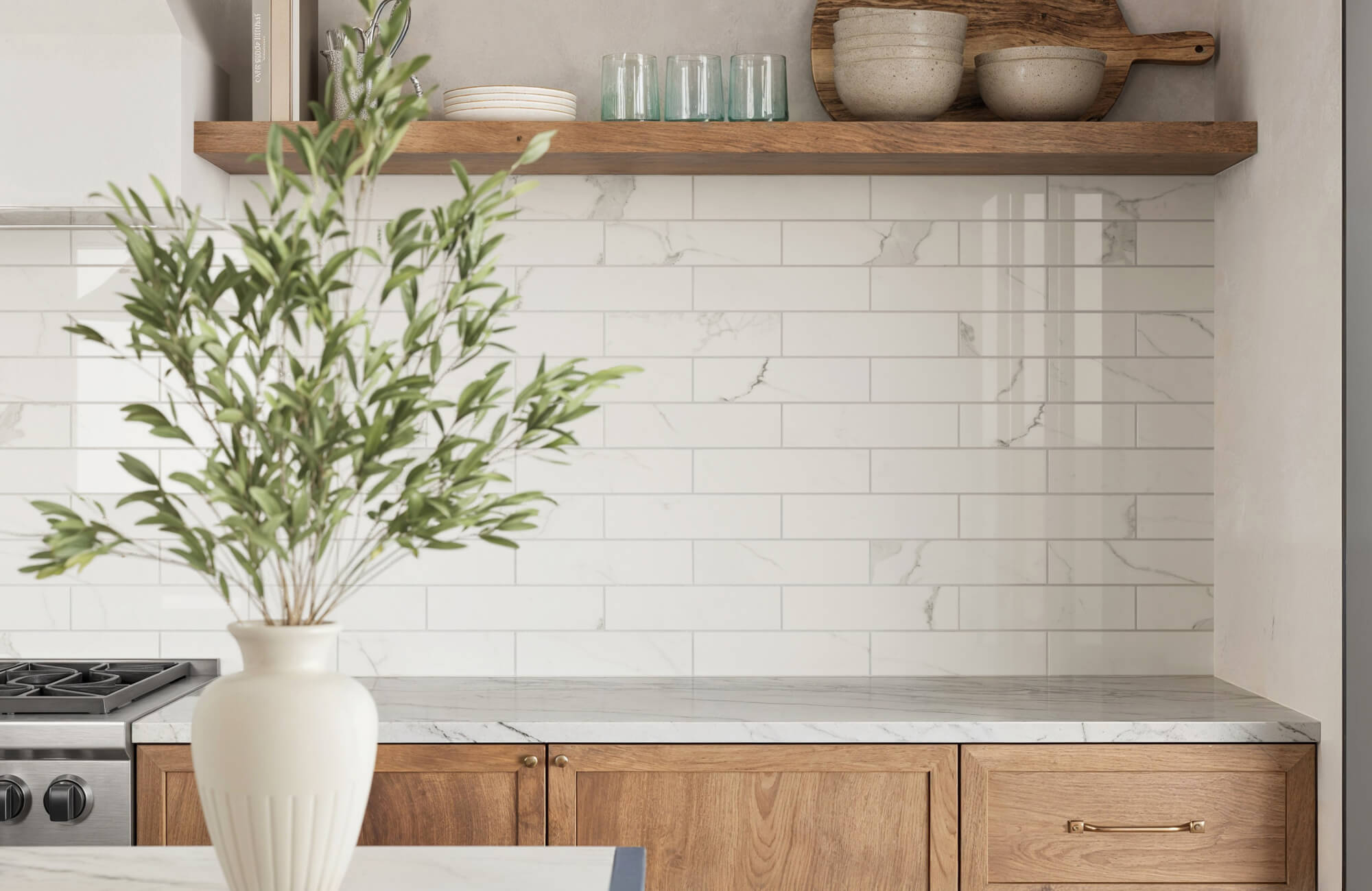 Modern kitchen with white marble-look subway tiles, wooden cabinetry, open shelving with rustic dishware, and a vase with greenery on the countertop.