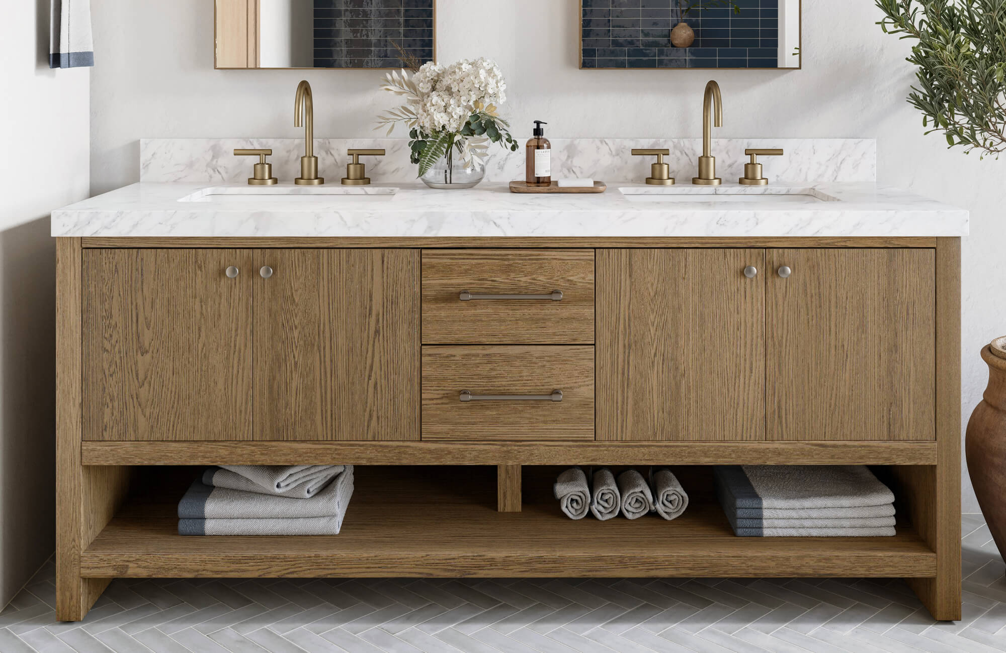 Warm oak double vanity with brushed brass faucets, marble countertop, and open shelving stocked with rolled towels.