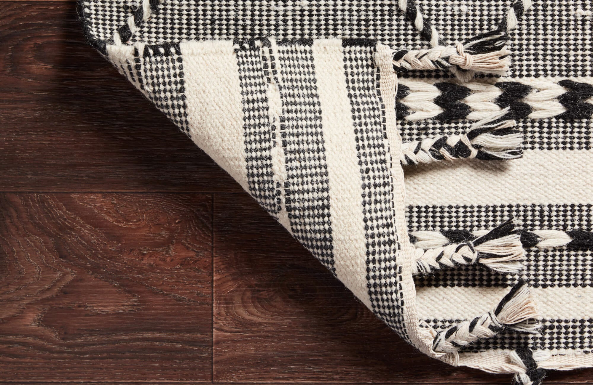 Black and cream high-quality woven rug with braided tassels and geometric stripes, folded over on rich walnut wood flooring.