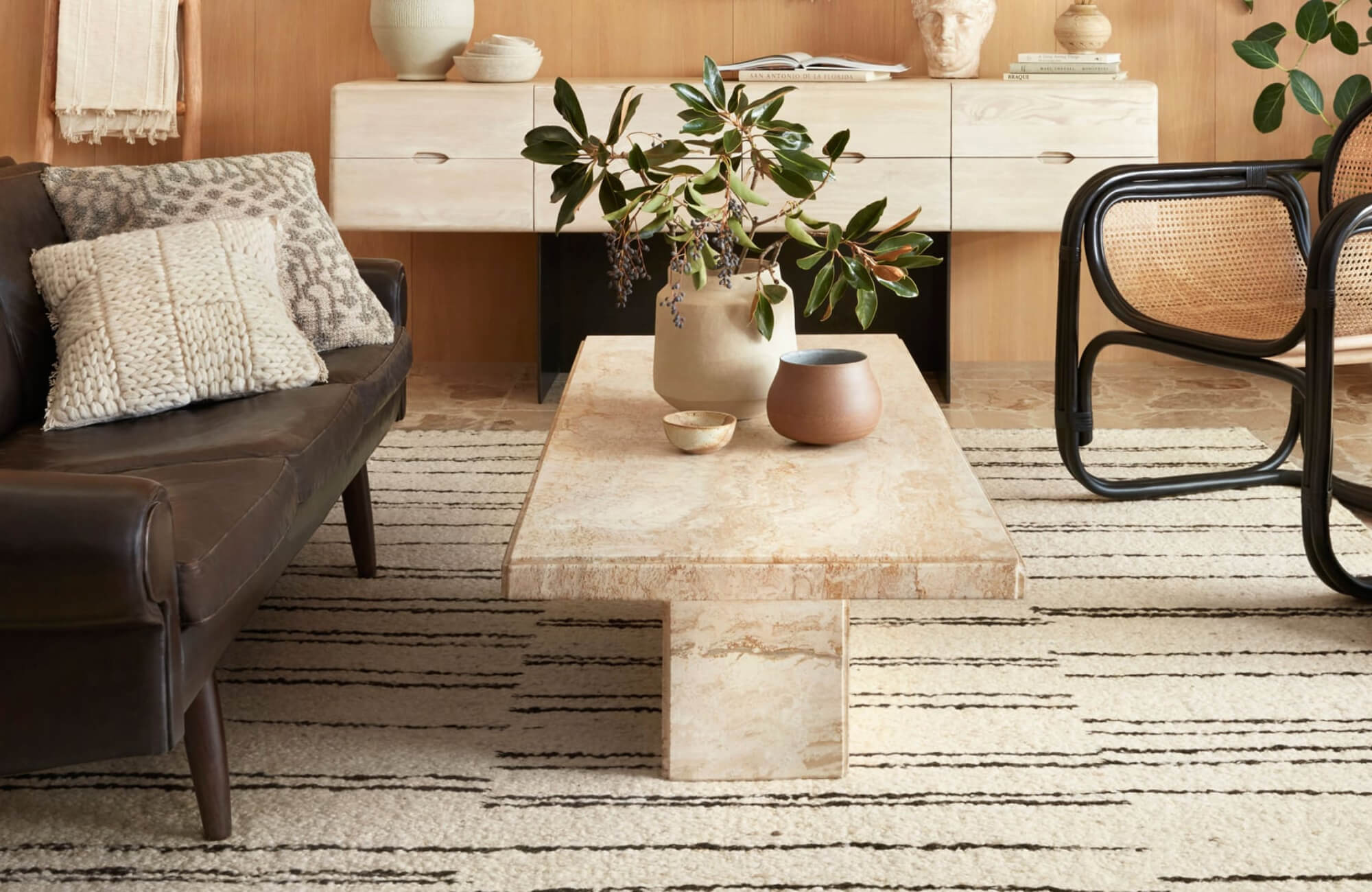 Warm-toned living room with a striped cream rug, stone coffee table, leather sofa, rattan chair, and minimalist decor accents.