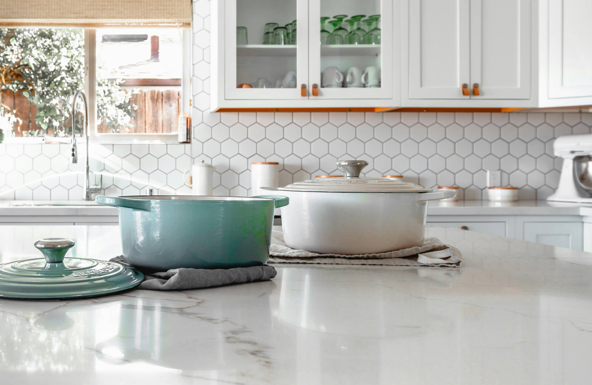 Bright kitchen with a white hexagon tile backsplash, marble countertops, and colorful cookware on the island.