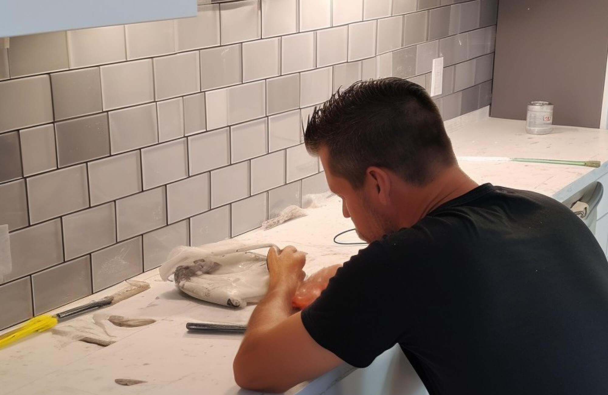 Close-up of backsplash installation in progress, featuring reflective gray ceramic tiles in a brick pattern above a white marble-like counter.