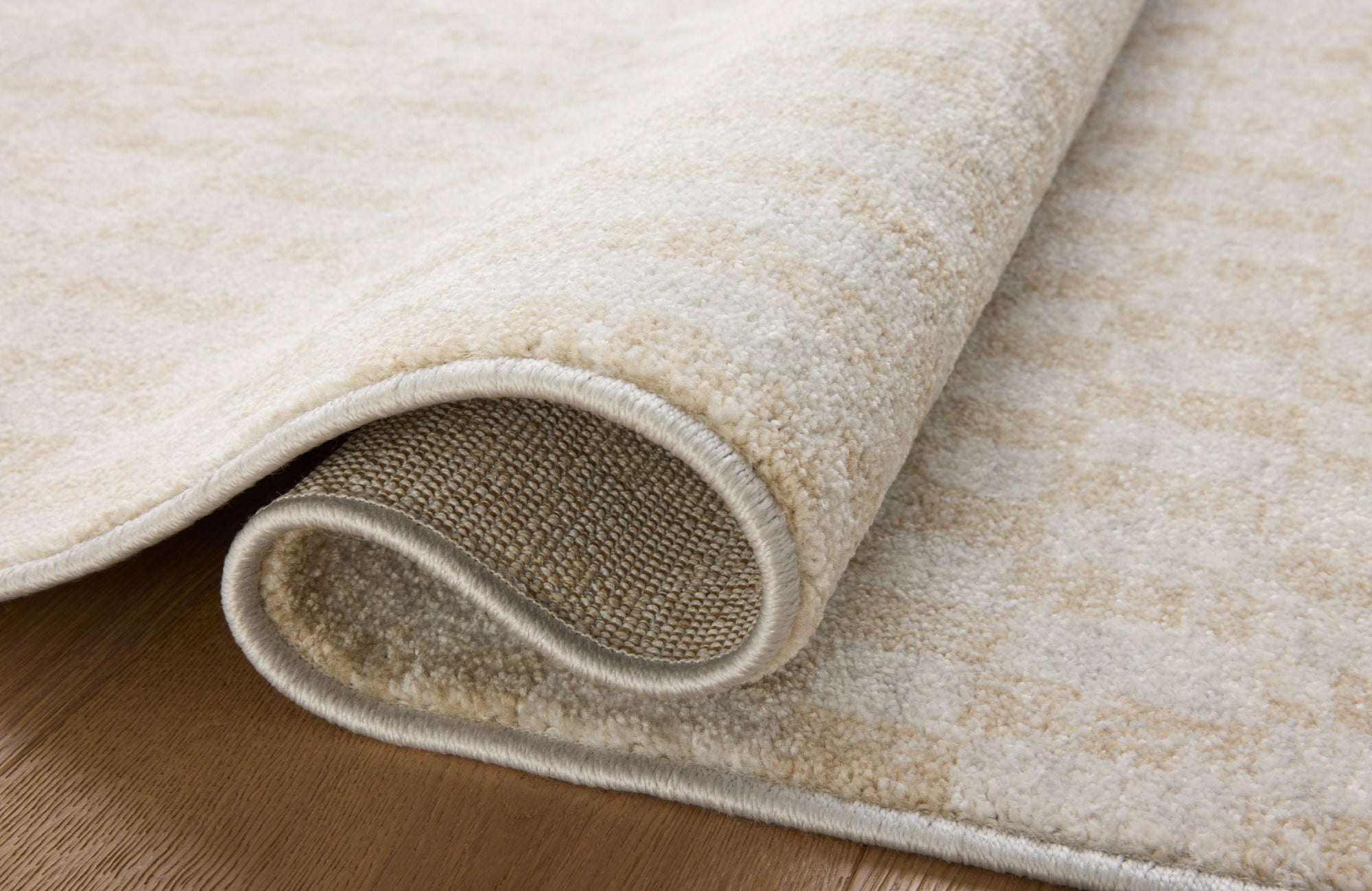 Close-up of a plush cream and sand polyester rug with a soft pile and neatly stitched edges, partially rolled on a wood floor.