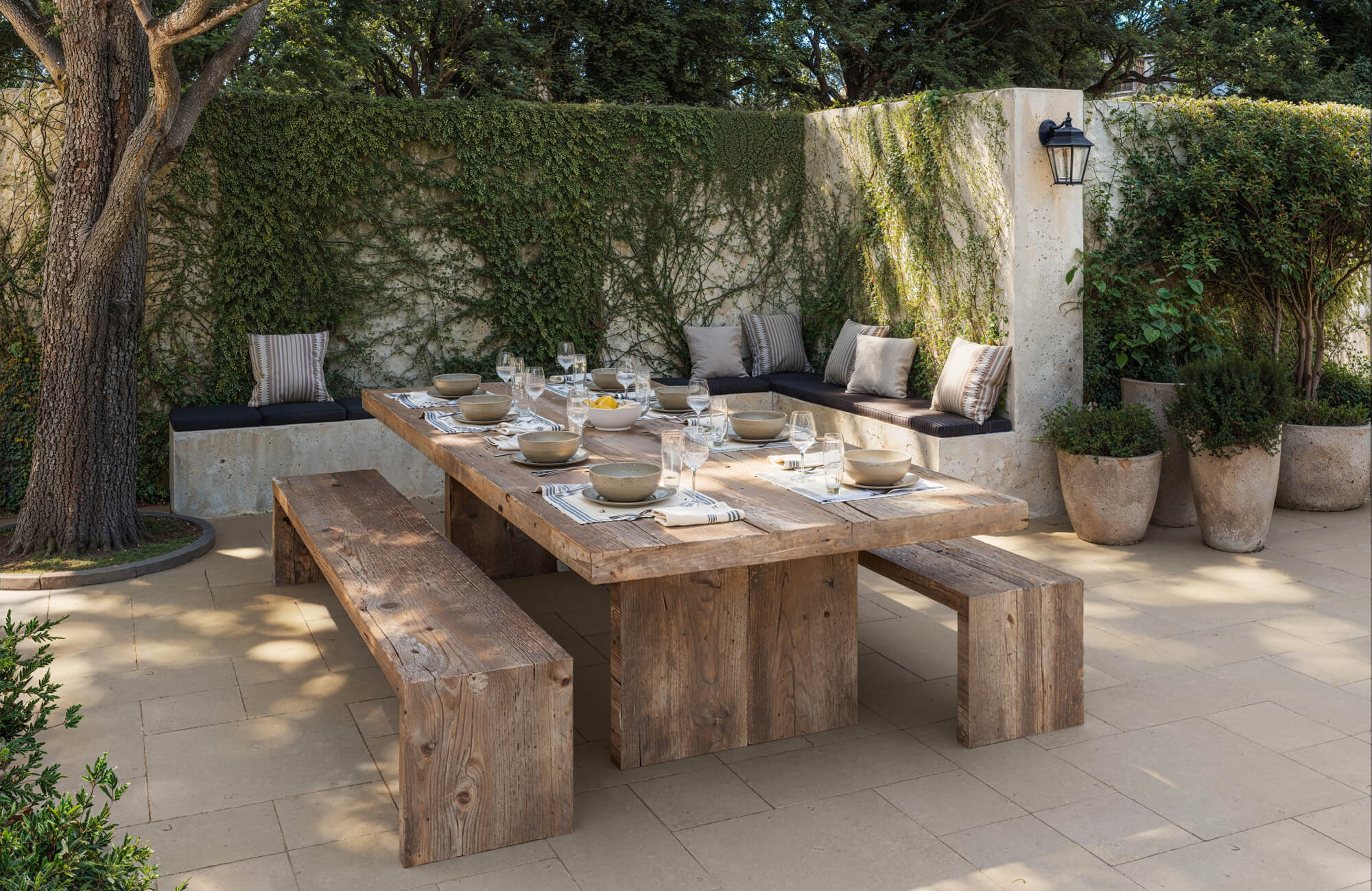 Charming outdoor dining area with warm beige porcelain pavers, rustic wood table set for guests, and vine-covered garden walls.