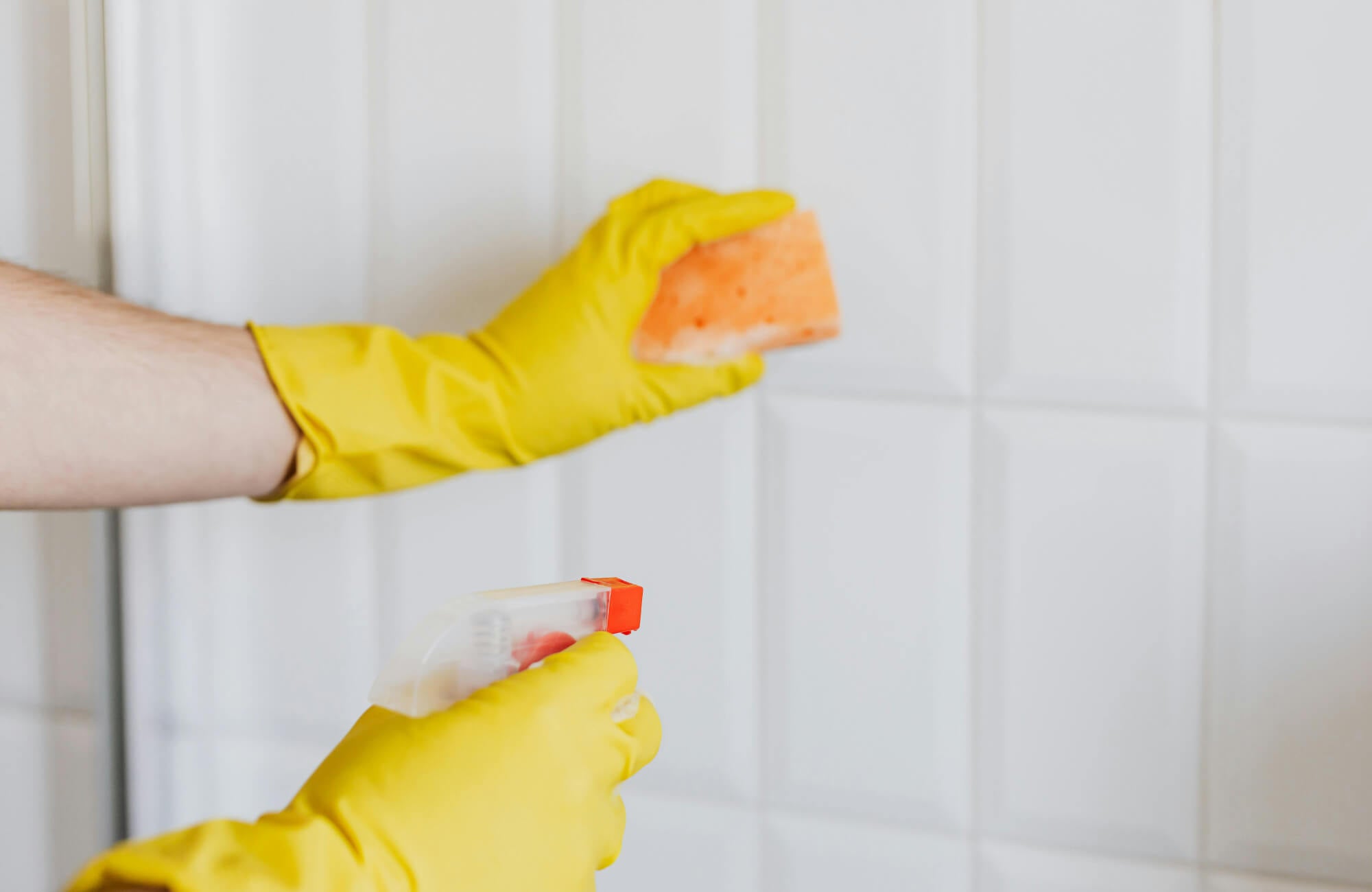 Gloved hands cleaning white textured ceramic tiles with a sponge and spray bottle for a fresh, polished finish.