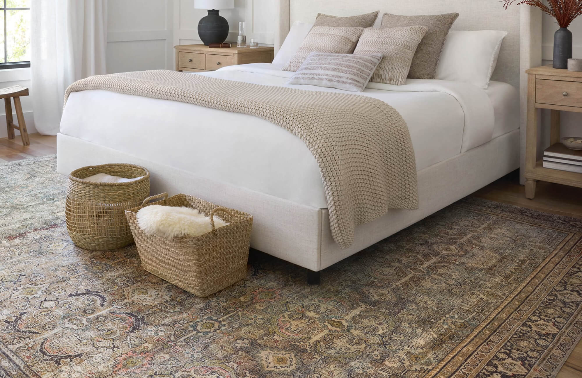 Cozy bedroom with an ivory upholstered bed, woven baskets, and a muted traditional rug in soft earthy tones.