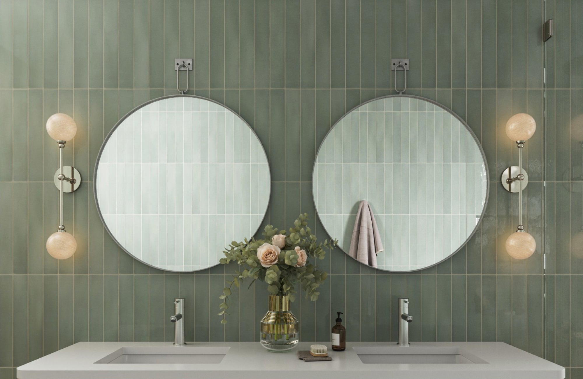 Pair of round mirrors in antique silver suspended by decorative mounts over a sage green tiled wall and vanity.