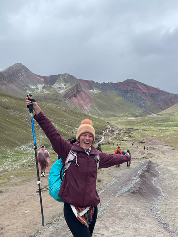 Ashley holding her arms and a hiking pole up in the air with green and red mountains in the background