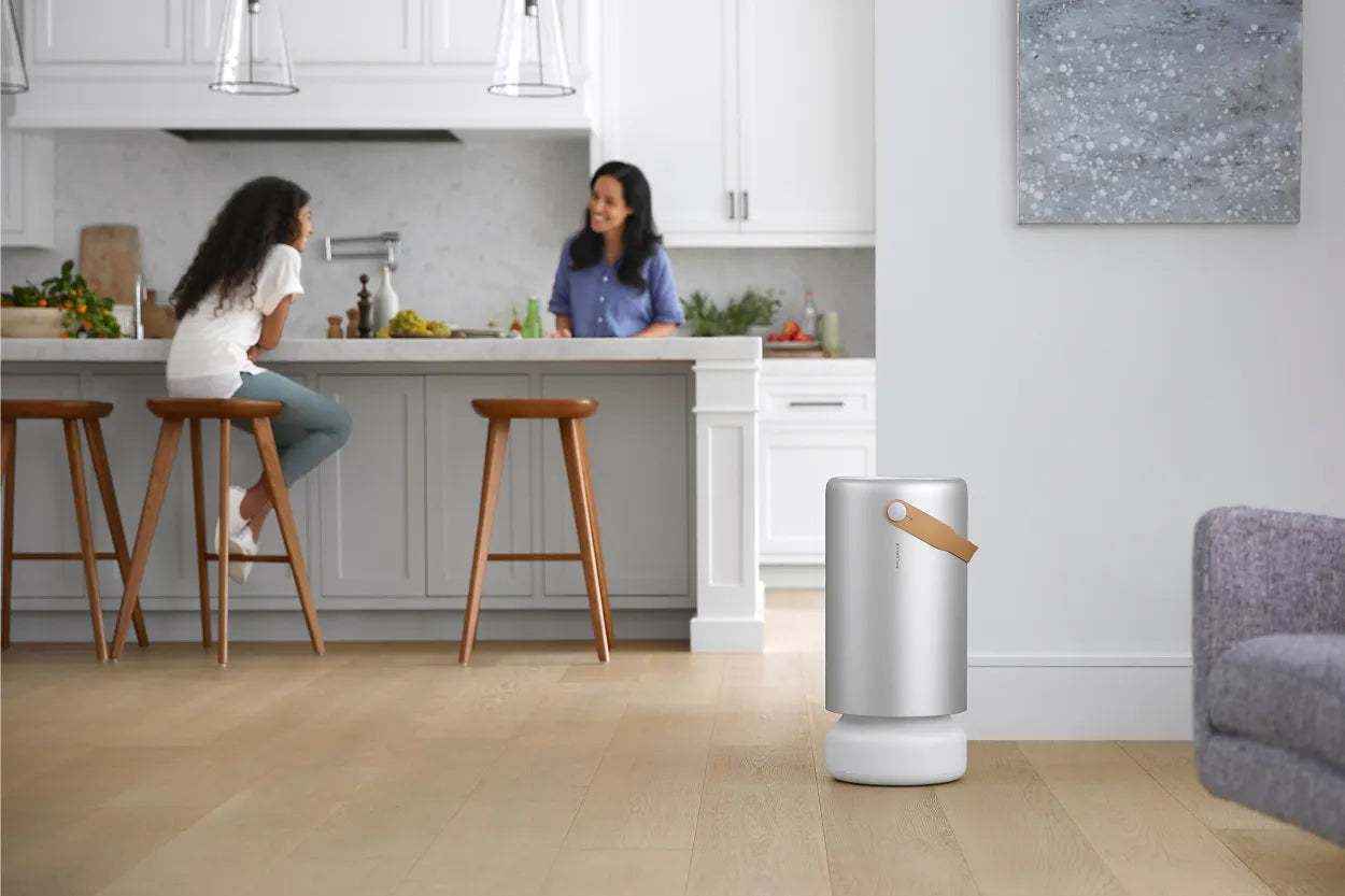 In the background, a mother and daughter sit around the kitchen island while an air purifier sits in the foreground of a modern-styled home.