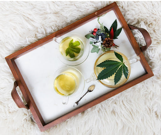 A tray on a fluffy rug with various cannabis leaves in pots and glasses to make a tea