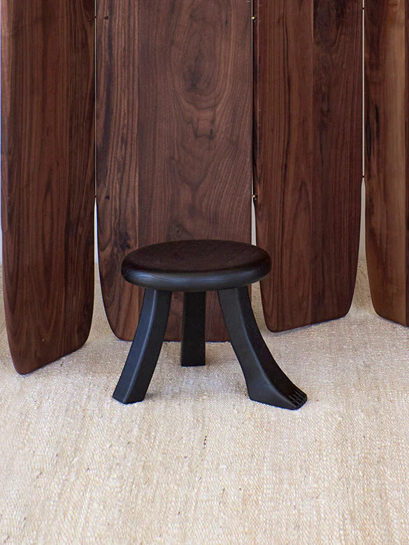 The Foot Stool in Black Chestnut by Project 213A, with its sculptural design, stands on a light rug. A room divider made from dark wooden panels enhances the elegance of the space, showcasing intricate wood grain.