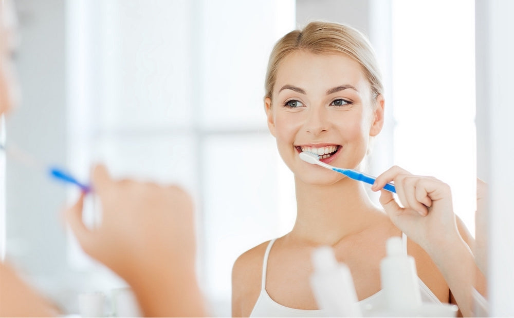 Afbeelding op de RichSmile-productpagina toont een vrouw die zichzelf van achteren in de spiegel bekijkt terwijl ze een tandenborstel vasthoudt. Ze glimlacht met sprankelende witte tanden die worden bereikt door RichSmile Serum te gebruiken.