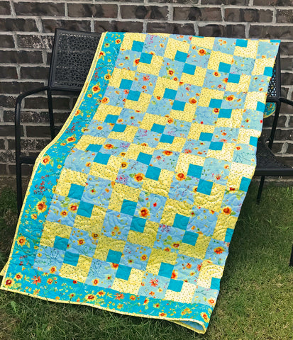 Cornerstones Quilt Pattern. Four patch patchwork quilt with teal and yellow floral fabrics. Quilt is shown displayed on a bench.