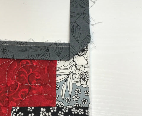 Red and gray patchwork quilt with gray binding being attached, showing the binding strip folded upward at 90 degree angle for mitered corner.