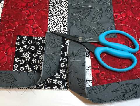 Red and gray patchwork quilt with two gray binding ends laying across it. Binding ends are both folded up at 90 degree angle and marked with white chalk along horizontal binding line. Scissors are shown nearby.