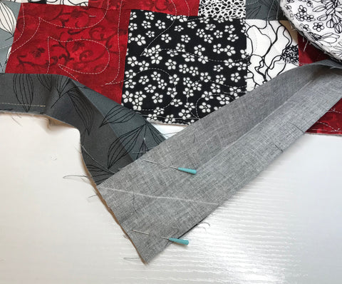 Red and gray patchwork quilt with two gray binding ends pulled downward and pinned at a 90 degree angle with white line drawn diagonally across them.