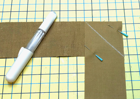 Two tan quilt binding strips pinned perpendicular to each other with a white chalk marker laying beside them.