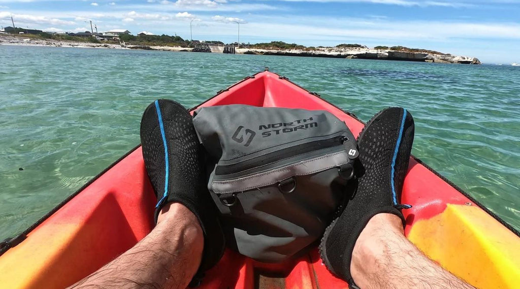 A mans feet at the end of a kayak with a North Storm Dry Bag in the front of the Kayak. 