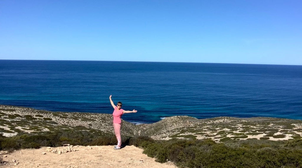 A lady standing on top of cliff face smiling with her arms outstretched. The ocean is behind her as she starts her adventures in Western Australia Gold detecting. 