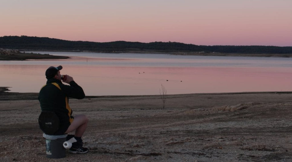 A MAN SITTING ON A BUCKET DOING A POOP BY A RIVER AND DRINKING A BEER WATCHING THE SUNSET.
