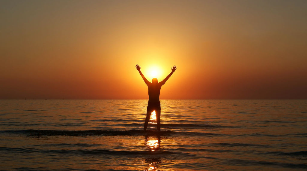 A MAN WITH HIS ARMS RAISED IN THE WATER AT SUNSET ON THE BEACH.
