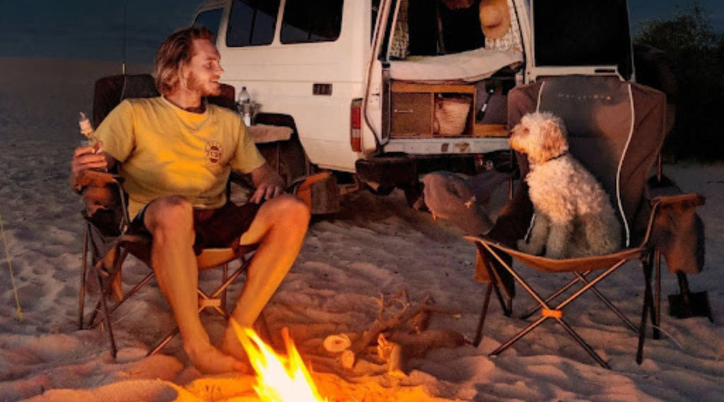 A MAN SITTING ON BEACH AROUND A CAMPFIRE LOOKING AND SMILING AT HIS DOG AT CORONATION BEACH CAMPGROUND, WESTERN AUSTRALIA, AUSTRALIA.