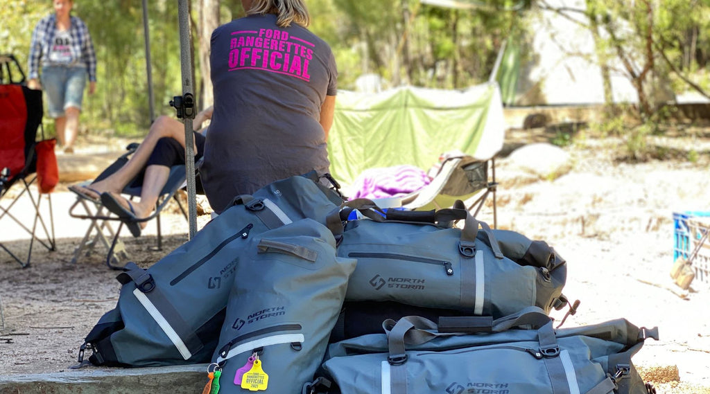 A PILE OF 4 - 5 NORTH STORM DUFFEL BAGS AT A CAMPSITE WITH A LADY SITTING IN FRONT WEARING A FORD RANGERETTES OFFICAL TSHIRT.