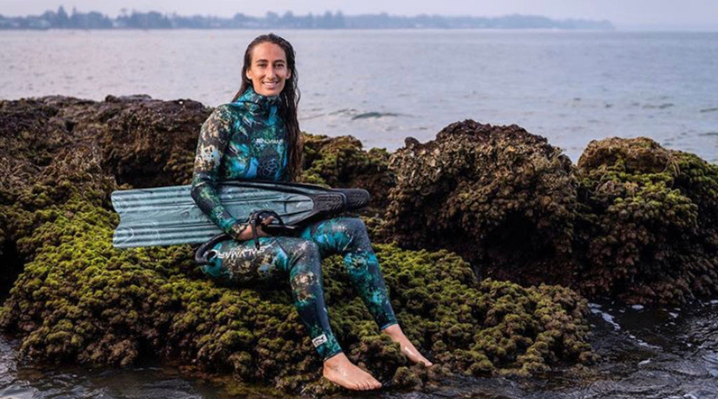 LARA FOUNDER OF WOEBEGONE FREEDIVE ON THE BEACH HOLDING FLIPPERS AND WEARING A WETSUIT