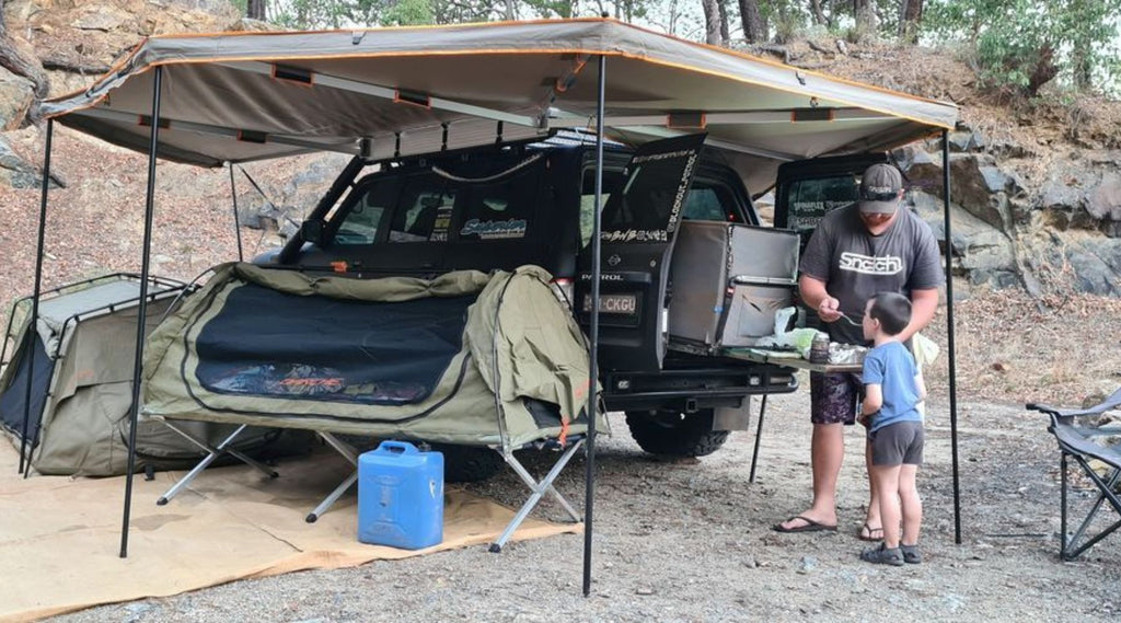 GAZ NORTH STORM AMBASSADOR FEEDING HIS SON AT A CAMPSITE STANDING UNDER AN TARP.