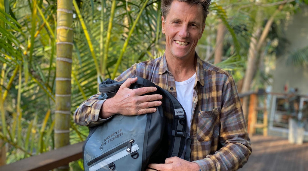 TIM BONYTHON HOLDING A NORTH STORM WATERPROOF BACKPACK AND SMILING.